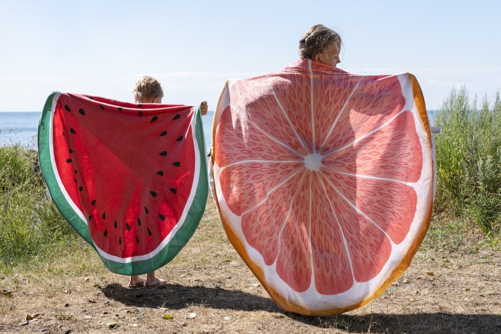 Två barn på stranden håller upp handdukar med frukttryck
